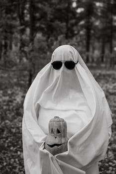 Woman in white sheet holding jack o'lantern against autumn forest background. Halloween costume. Black and white photo