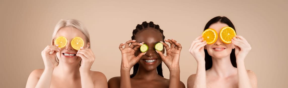 Beauty And Skin Care. Three Diverse Women In Bath Towels Covering Eyes With Cucumber And Orange Slices, Making Organic Face Masks, Posing Together Over Beige Studio Background, Copy Space