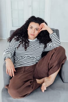 Curly brunette woman in homemade coat sitting on armchair smiling