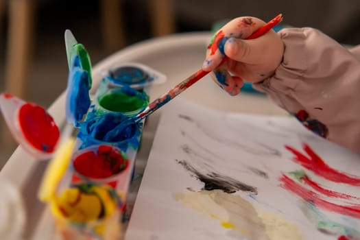 Close up of a toddlers hand reaching for a paintbrush with gouache colors. Creative activity at home, early childhood art education.