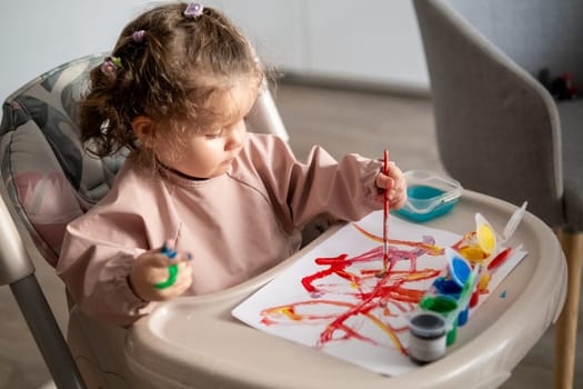Cute toddler girl painting with gouache at a table near a sunlit window. Natural light highlights her artwork and the joyful atmosphere of creativity.