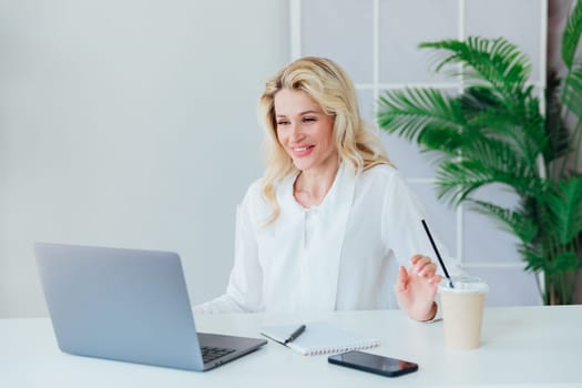 Blonde woman working on laptop and smiling at desk in office online