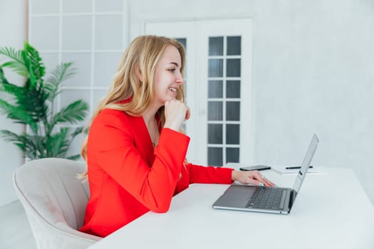 Business blonde woman in red suit at desk with laptop working in office