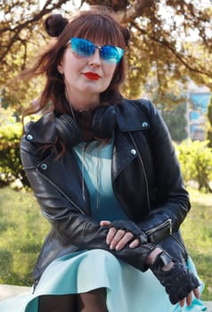 Attractive girl in black leather jacket, blue dress., black gloves, blue sunglasses looks into the camera.