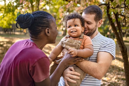 Two parents embrace their joyful baby boy in a vibrant park. Sunlight filters through colorful trees, creating a warm, loving atmosphere as laughter fills the air.