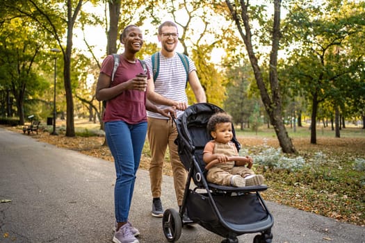 A couple enjoys a leisurely walk in a vibrant park, surrounded by autumn leaves. Their playful baby in a stroller observes the beautiful day, creating a warm family moment.