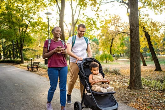 A joyful couple walks hand in hand through a picturesque park, enjoying a sunny autumn day while pushing a stroller with a curious child. Leaves create a vibrant backdrop.