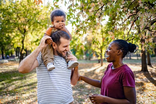 A cheerful family enjoys a sunny day in a lush park, surrounded by colorful fall foliage. The father carries his smiling child on his shoulders while the mother shares laughter nearby.