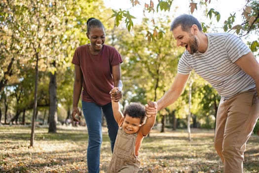 A cheerful family enjoys a sunny day at the park, as a child giggles joyfully while being lifted by both parents. The green trees create a vibrant backdrop for their playful bonding.