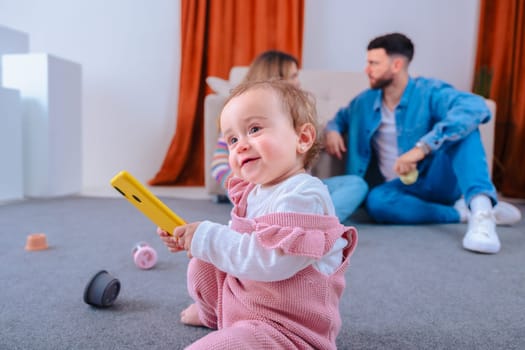 Playful baby daughter in pink overalls sits on carpet gripping a yellow smartphone, exploring technology, while her calm parents chat on the sofa in a living room,capturing daily family life at home