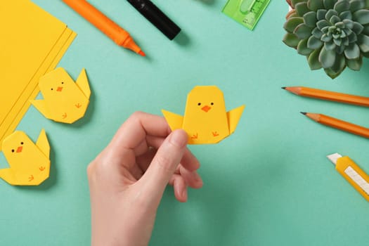 Making a simple Easter origami handicraft. Holds in his hand a ready-made paper craft of a chicken. Around the tools for crafts