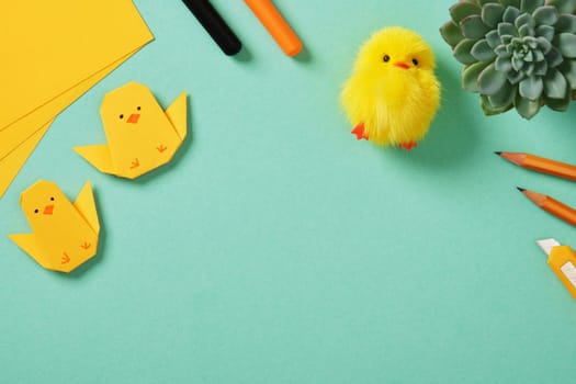 Frame on turquoise background with two origami chicks made of yellow paper, fluffy yellow toy chick. Art supplies: pencils, markers, drawing knife. Place for text