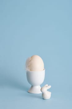 Minimalist Easter egg with a white feather bird and ceramic bunny on a pastel blue background. A place for your text, congratulations, wishes.