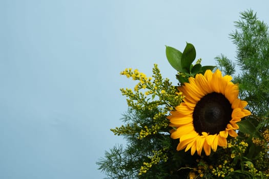 Brightly colored bouquet with yellow sunflower and green foliage on a blue background. Place for your text.