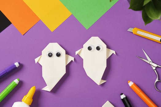 Origami paper ghosts with googly eyes surrounded by colorful craft supplies on a purple background. Paper sheets, scissors, markers, glue and knife for children's crafts or holiday DIY projects.