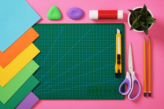 Supplies for paper art hobby: cutting mat, scissors, craft knife, pencil, eraser, glue colored paper on pink background. Place for your text. Paper crafts work table flatlay.