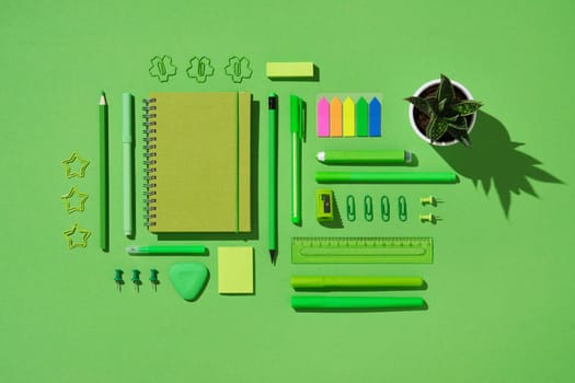 Flat lay of green stationery set including notebook, pens, pencil, ruler, sticky notes, paper clips, push pins, eraser, and small potted plant on bright green background
