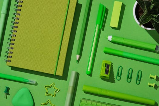 Close-up flat lay of assorted green office supplies on vibrant background, featuring spiral notebook, pens, pencil, sharpener, sticky notes, eraser, paper clips, push pins, and a small potted plant
