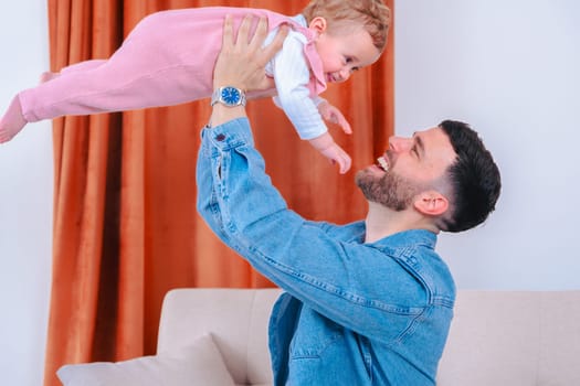 Young father in denim jacket joyfully raises his smiling baby daughter in pink overalls high above a sofa in a living room,capturing playful bonding, trust, love and shared family happiness together