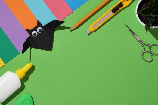 Creative workstation with colorful craft materials on a green background with a handmade black origami bat with googly eyes, scissors, utility knife, glue bottle, eraser, pencil and colorful paper sheets.