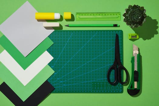 Top view of the paper crafts work table in green theme. Scissors, craft knife, pencil, eraser, cutting mat, colored paper. Space for text.