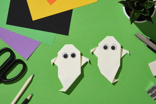 Two white paper handmade origami ghosts with googly eyes are arranged on a green background surrounded by colorful sheets, pencils, scissors, cutter, eraser and sharpener.