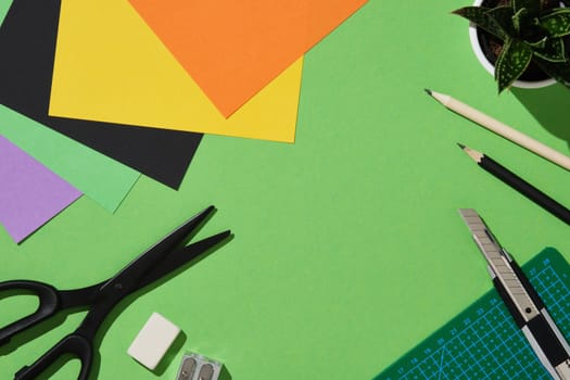 Frame of supplies for paper art hobby: scissors, craft knife, pencil, eraser, cutting mat, colored paper on green background. Copyspace.