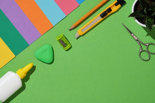 Tools for paper art hobby: scissors, craft knife, pencil, eraser, cutting mat, colored paper on green background. Copyspace. Paper crafts work table flatlay.