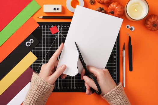 Seasonal diy paper crafting. Hands cut a ghost silhouette out of white paper with scissors. Around paper crafting items, pencil, tape, cutter, tweezers.