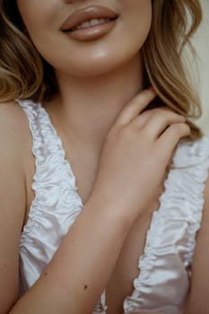 cropped close-up of attractive blonde in white silk lingerie. Lips in frame and hand. wavy hair, beautiful makeup. close-up portrait. Concept of face and skin care at home, bride's morning