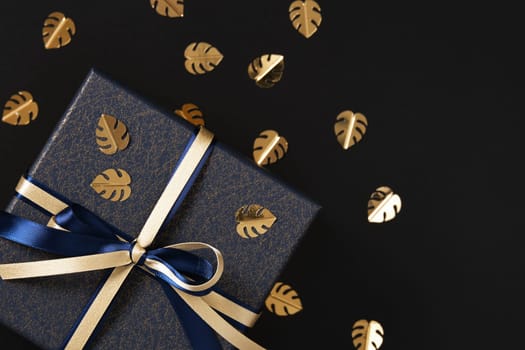 Fragment of blue gift box with gold and blue satin ribbon. Black background, gold confetti in the form of monstera leaf, space for text.