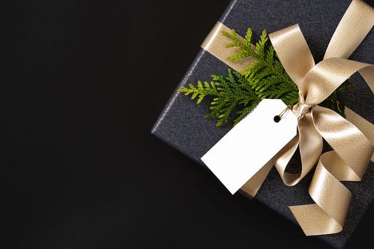 Blank gift tag on blue gift box with gold satin ribbon and tui sprig. Black background, space for your text.