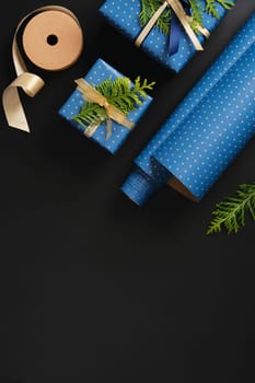Flat composition with blue gifts with gold ribbons, brown paper tags, green thuja branches on black background. Space for your text.