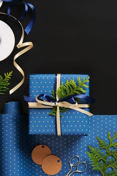 Flat composition with blue gifts with gold ribbons, brown paper tags, green thuja branches on black background. Space for your text.