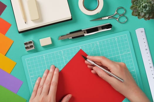 Hands working with colorful paper using a precision knife on a mint green cutting mat. Tools for paper crafts. Ruler, pencil, scissors, tape, eraser