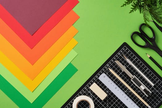 Neatly arranged paper craft supplies. Black cutting mat, cutter, scissors, ruler, pencil, tape, colored paper. Green background, space for your text.