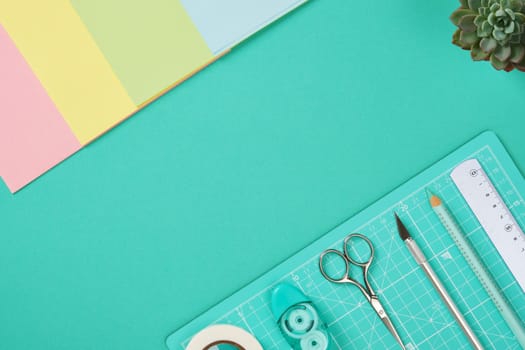 Neatly arranged materials for paper crafts. Mint-colored cutting mat, cutter, scissors, ruler, pencil, tape, colored paper. Turquoise background, space for your text.