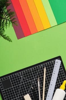 Top view of a desk for paper crafts. Black cutting mat, craft knife, scissors, ruler, pencil, glue, colored paper. Green background, space for your text.