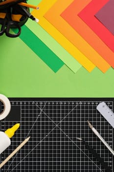 Top view of a desk for paper crafts. Black cutting mat, craft knife, scissors, ruler, pencil, glue, tape, colored paper. Green background, space for your text.