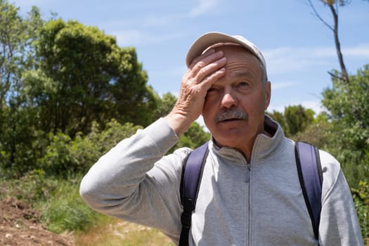 An elderly man suffering from the heat while walking in the forest, wiping sweat with his hand. High quality photo