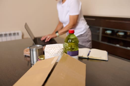 A woman is seen organizing a food package with pantry essentials like oil and canned goods while working on her laptop, suggesting donation or online shopping.