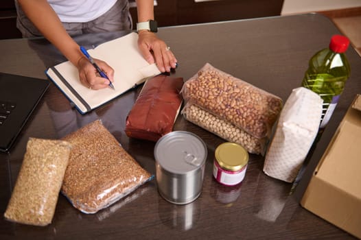 A person arranges groceries like canned goods, beans, and grains on a table, recording details on a notepad, depicting organization and planning for sustainable living.