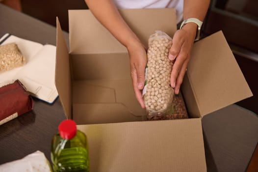 Individual organizing pantry items, including beans and bottles, in a cardboard box, emphasizing assistance, support, and food preparation during daily tasks or charitable activities.