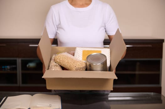 A woman in a white T-shirt holding a box containing food staples including canned goods, grains, and packaged items. The box rests on a table with a notebook next to it.
