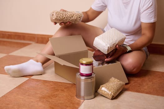 Close-up woman places food items into a box, preparing a donation. The scene depicts a mindful act of helping by organizing essentials in a home setting. Food bank Day and Humanitarian aid concept