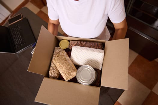 A close-up of a person carrying a cardboard box filled with non-perishable food and essentials in a home setting with a table in the background.