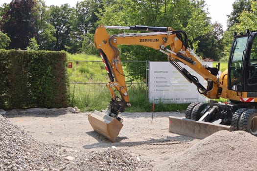 09.06.2025 Germany: Excavator at construction site during landscaping work. Yellow excavator with the brand name ZEPPELIN working on a construction site surrounded by gravel piles and greenery. A banner with tree care and soil services is visible in the background, indicating landscaping or land preparation work in a rural or suburban area.