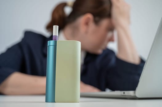 Tired businesswoman smoking an electronic cigarette while working on a laptop. Tobacco heating system