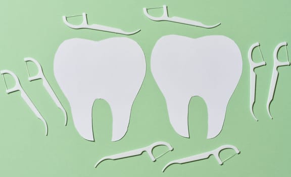 Two white paper cutouts of teeth are surrounded by multiple white dental floss picks on a light green background, emphasizing dental hygiene