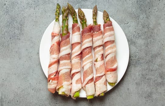 Delicious asparagus wrapped in bacon strips, seasoned with colorful peppercorns, presented on a white plate, ready to be cooked
